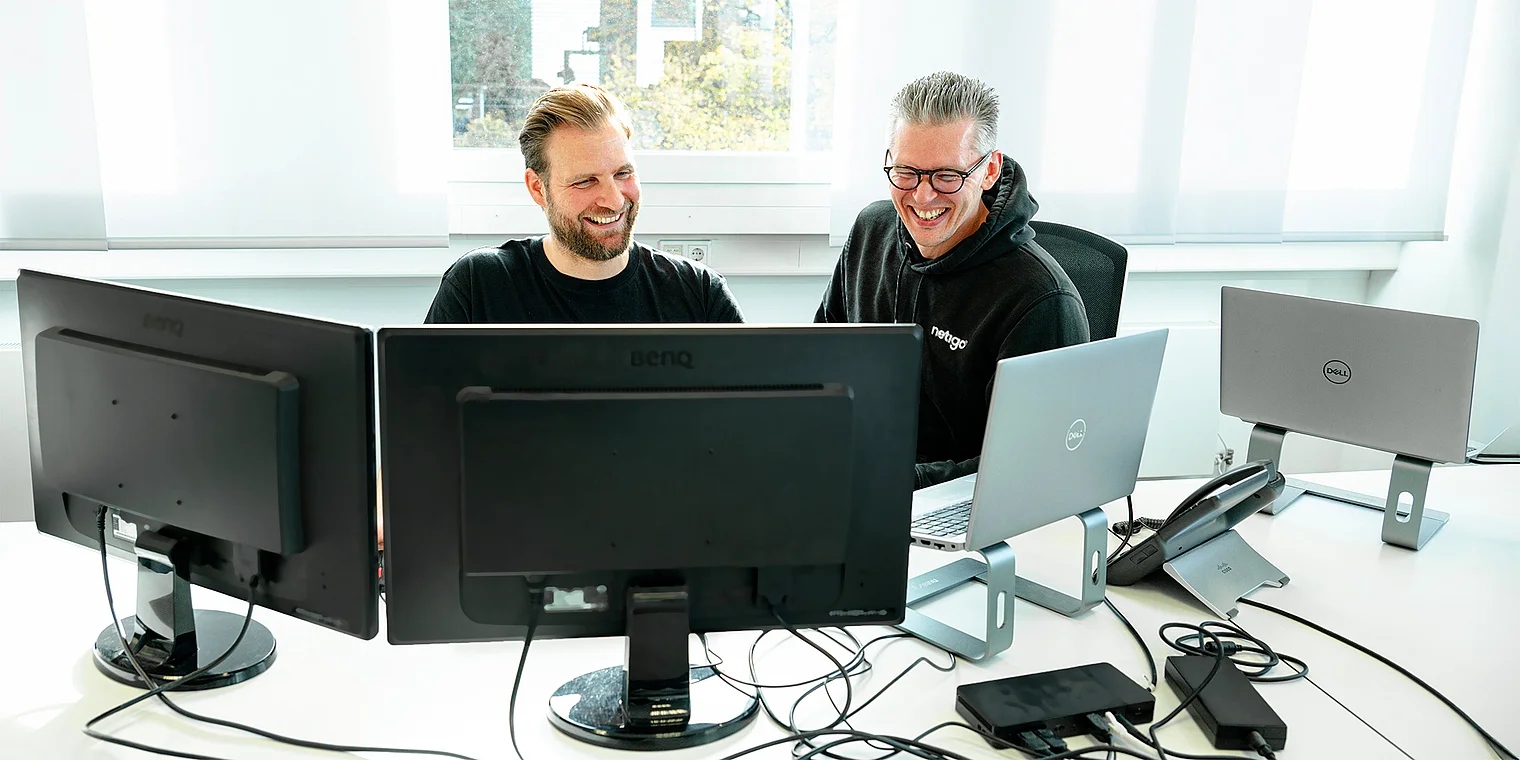 Sebastian und Walter, beide Webdesigner/Entwickler bei der Webagentur Netigo, stehen nebeneinander und lächeln in die Kamera; das Bild visualisiert die Webdesign-Leistungen der Agentur mit einem thematisch passenden Hintergrund, der digitale Gestaltung und kreative Nutzeroberflächen symbolisiert.