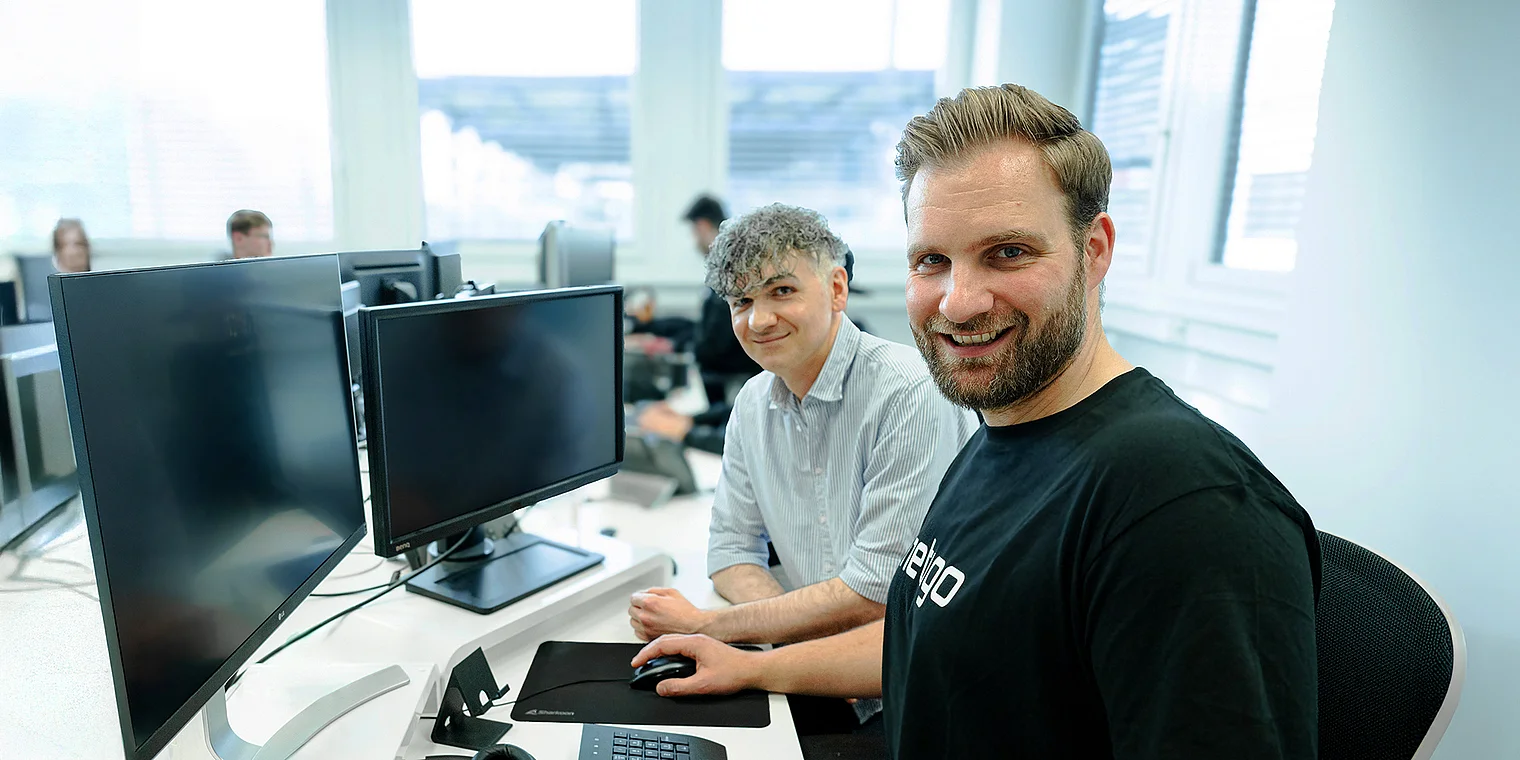 Sebastian und Kemal, beide Entwickler bei der Webagentur Netigo, stehen nebeneinander und lächeln in die Kamera; das Bild visualisiert die Programmier-Leistungen der Agentur, mit thematisch passendem Hintergrund für digitale Entwicklung und technisches Arbeiten.